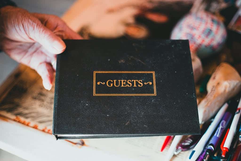 A hand holds a guest book surrounded by pens and a blurry background.